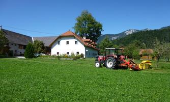 A farmhouse with a red tractor on a green meadow. In the background, gentle hills and a clear blue sky can be seen.