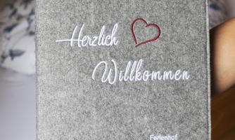 A gray welcome book with the inscription "Herzlich Willkommen" and a heart. It lies on a table in a cozy room.