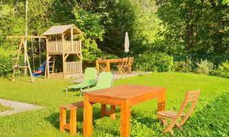 A beautiful garden with a wooden table and colorful chairs. In the background, there is a playhouse and a swing.