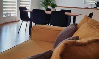 A modern living-dining room with a comfortable sofa and a dining area. Large windows with blinds let in plenty of light.