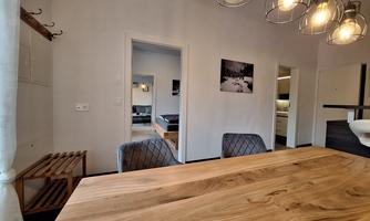 A stylish dining area with a large wooden table and modern chairs. In the background, additional rooms can be seen that appear bright and inviting.