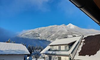 A snowy winter landscape with a clear blue sky and mountains in the background. The buildings in the foreground are covered with snow.