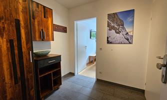 A bright foyer with a wooden cabinet and a modern picture on the wall. On the side, an open door can be seen, leading into another room.
