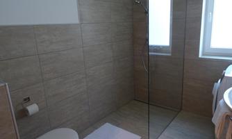 A modern bathroom with a shower and a toilet. The walls are adorned with light tiles and there is a window for daylight.