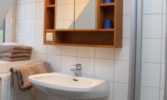 A modern bathroom with a white sink and a wooden shelf. Towels are neatly stacked and the mirror reflects the room.
