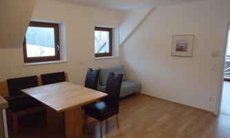A cozy living space with a wooden table and black chairs. Bright walls and large windows provide ample light.