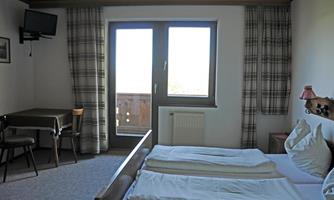 A cozy guest room with two beds, a table, and chairs. Large windows provide plenty of daylight and a view of the balcony.