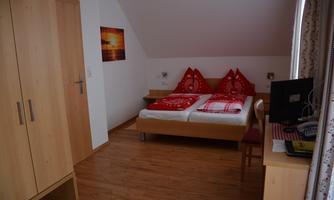 A simple bedroom with two beds, red pillows, and a desk. A picture hangs on the wall, and there is a wooden wardrobe.
