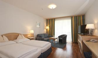 A comfortable hotel room with two beds, a sofa, and an armchair. Large windows provide plenty of daylight.