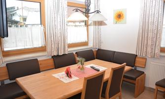 A cozy dining area with a large wooden table and chairs. Windows with curtains let in a lot of light.