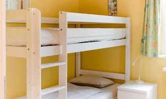 A cozy children's room with a bunk bed and white furniture. The walls are painted in a warm yellow tone and a window lets in plenty of light.