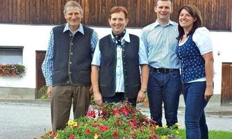 A group of four people stands smiling in front of a house, surrounded by colorful flowers. They are wearing traditional clothing and appear friendly and welcoming.