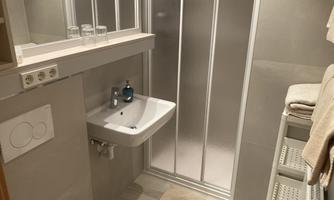 A modern bathroom with shower, sink, and towels. The walls are tiled in light gray, and there are practical shelves.