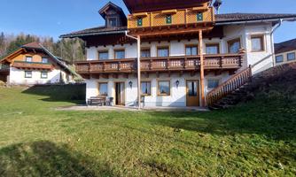 A picturesque wooden house with multiple floors and a large balcony. The garden is green and well-maintained, surrounded by trees.