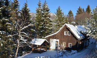 A charming wooden cabin in the snow, surrounded by tall fir trees. The clear blue sky gives the winter landscape a peaceful atmosphere.