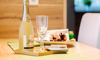 A bottle of sparkling wine stands on a wooden board next to a glass goblet. In the background, some decorative elements and a green tablecloth are visible.