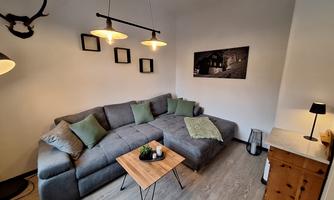A cozy living room with a gray sofa, green cushions, and a small wooden table. Picture frames hang on the wall, and a decorative lamp provides warm light.