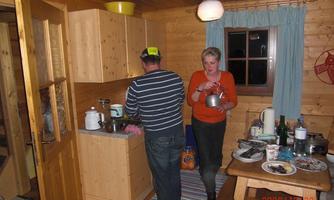 Two people in a cozy wooden kitchen. One person is standing at the sink and the other is carrying a kettle.
