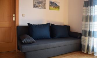A cozy sofa with blue cushions in a bright room. On the wall hang two pictures, and a window with curtains provides light.