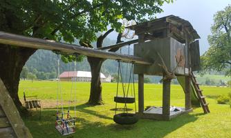 A playground with a treehouse structure and swings. In the background, meadows and trees can be seen.