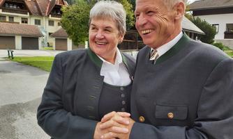 A smiling couple in traditional attire stands close to each other. They are holding hands and appear happy.
