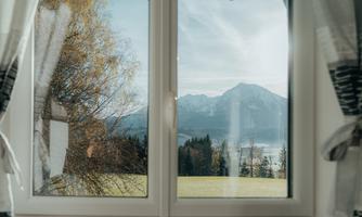 View from a window of a mountain landscape with trees and meadows. The sky is bright and partly cloudy.