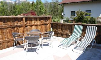 A sunny terrace with a table and six chairs, as well as two loungers. In the background, green trees and a residential house can be seen.