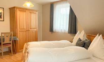 A cozy bedroom with two wooden beds and white bedspreads. In the room, there is a wardrobe and a small table.
