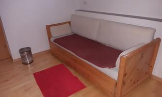 A wooden sofa with a light covering and a red throw. Next to it lies a red rug piece on a light wooden floor.