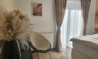 A cozy bedroom with a large bed and a chair. The windows are decorated with light curtains and there is a decorative plant element on the table.