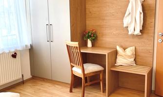 A bright room with wooden flooring and a desk. On the wall, there is a piece of clothing and a small vase with flowers.