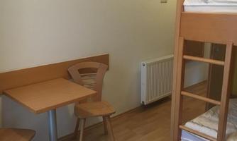 A simple room with a loft bed, a table, and two chairs. The room has a bright, neutral color tone and a radiator.