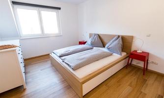 A bright bedroom with a double bed and two large pillows. Next to the bed is a red nightstand and the floor is wooden.