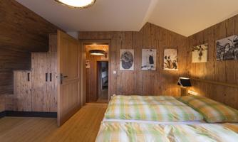 A cozy bedroom with wooden panels and a double bed dressed in striped bedding. Pictures hang on the walls, and there is a staircase leading to another room.