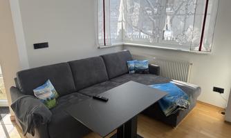 A cozy living room with a gray couch and a small table. The windows are decorated with light curtains and let in a lot of daylight.