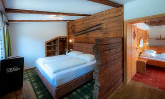 A cozy room with a large double bed and rustic wooden walls. There is a small table and a connection to another sleeping area.