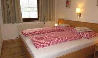 A cozy bedroom with a large double bed and checked blankets. The room has a window with curtains and gentle wall lighting.