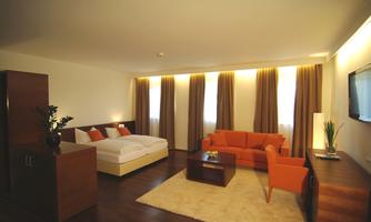 A modern hotel room with a large bed, a cozy sofa, and wooden flooring. The walls are painted in light colors and large windows let in plenty of light.