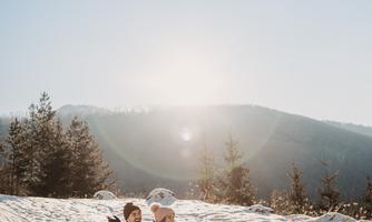 A couple sits on a sled in the snow, enjoying the sunshine. In the background, snow-covered hills and a blue sky can be seen.