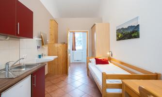 A simple room with a single bed, a small kitchenette, and a table. The room has bright walls and large windows that let in natural light.