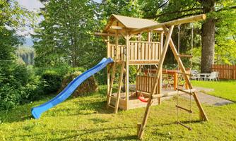 A playground with a wooden play castle, a slide, and a swing. The surroundings are green and provide space for playing.