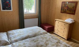 A cozy bedroom with two single beds and wood paneling. The room features a wardrobe, a stool, and artwork on the walls.