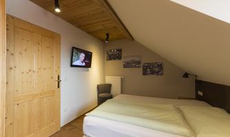 A cozy room with a double bed and a television on the wall. The wooden door and ceiling give the space a warm character.