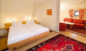 A cozy bedroom with two single beds and light wood. In the background, there is a seating area with a red couch and an oriental rug.