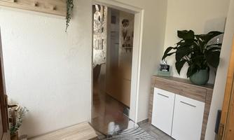 A bright hallway with a wooden shelf and a plant. On the left side, there is a bench and a cabinet with white doors.