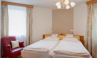 A bright bedroom with two single beds and a cozy red armchair. Large windows provide ample daylight.