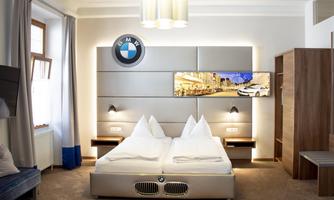 A modern hotel room with two beds and a BMW design wall. A television shows a scene of a city.