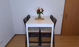 A modern dining area with a white table and four chairs. In the center of the table is a vase with fresh flowers.