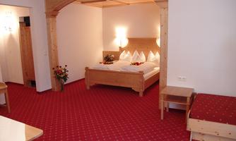 A cozy bedroom with a large double bed and decorative pillows. The floor is covered with a red carpet, and there is a small side table.