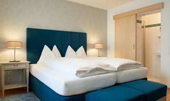 A cozy room with a large double bed and blue accents. There are nightstands and a shower in the background.